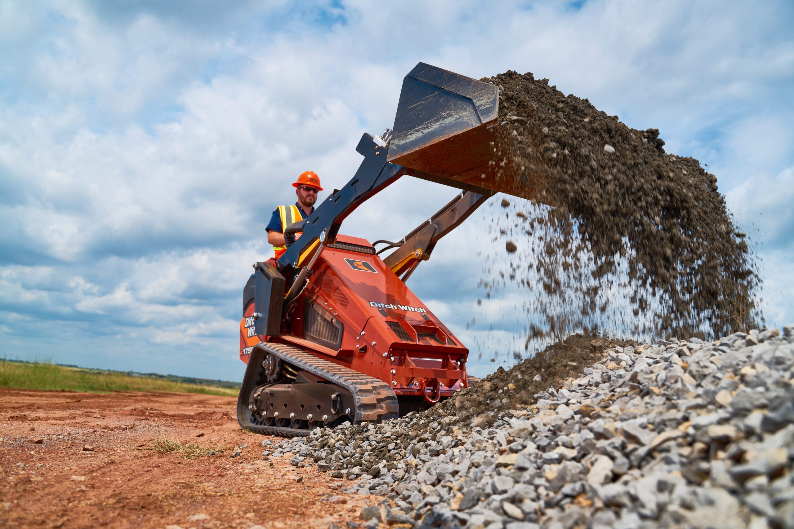 Ditch Witch SK1750 skid steer loader dumping gravel at a construction site using a front loader bucket.