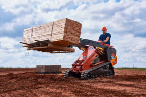 Ditch Witch SK1750 stand-on skid steer lifting a pallet of lumber with pallet forks on an active jobsite.