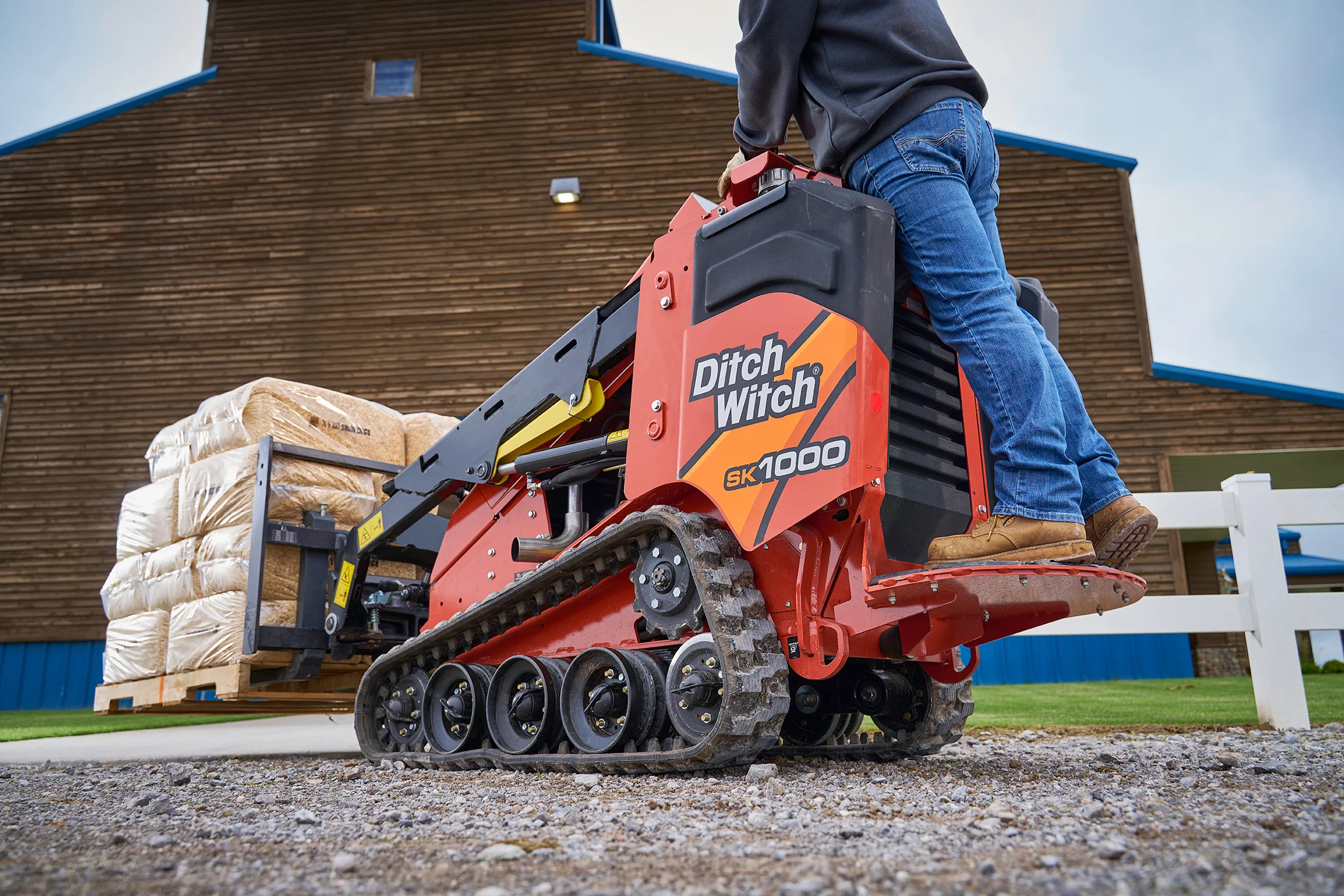 Ditch Witch SK1000 mini skid steer using a pallet fork attachment to transport a pallet of bagged materials outside a building.